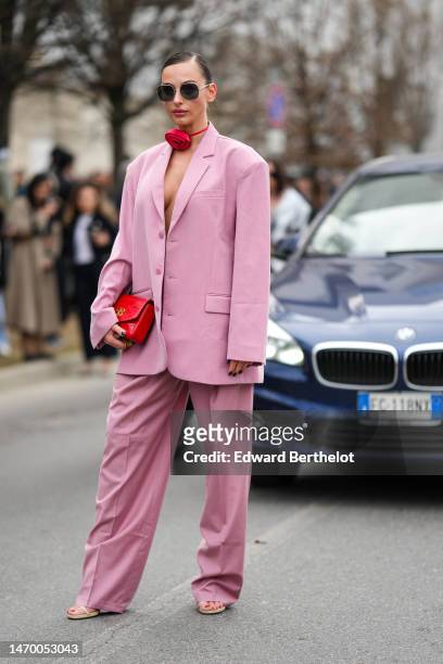 Guest wears sunglasses, a red silk flower necklace, a pale pink oversized blazer jacket, matching pale pink large suit pants, a red shiny leather...