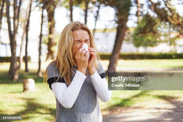 woman suffering from spring allergy, blowing nose with a tissue in the park - sinusitis stockfoto's en -beelden