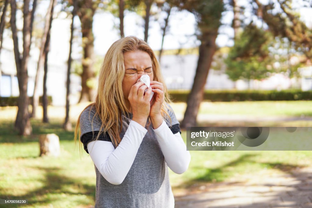 Woman suffering from spring allergy, blowing nose with a tissue in the park