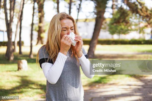 woman suffering from spring allergy, blowing nose with a tissue in the park - sneezing stock pictures, royalty-free photos & images