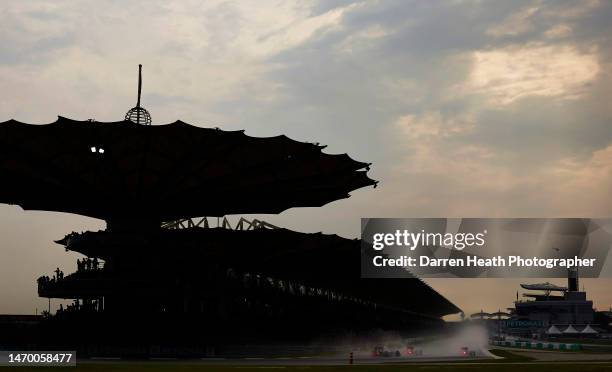 Under a cloudscape sky, Formula One team cars driving past the start and finish straight stadium grandstands in wet, spray inducing rain conditions...