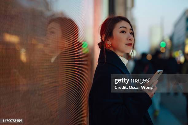 young asian business woman using smart phone against led lights on city street - high street bank uk stock pictures, royalty-free photos & images