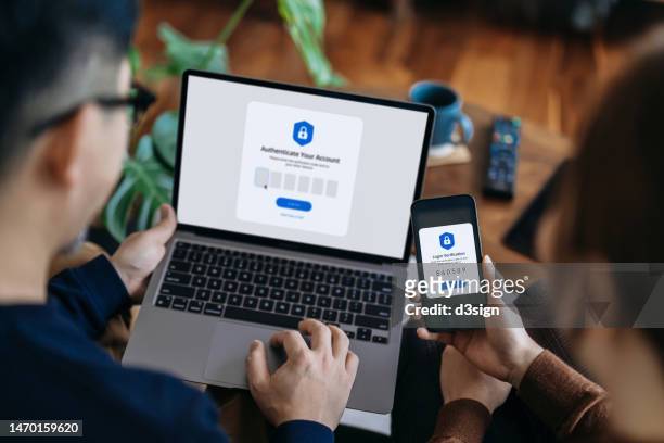 over the shoulder view of young asian couple using mobile device with two-factor authentication (2fa) security while logging in securely to their laptop at home. privacy protection, internet and mobile security concept - código de segurança - fotografias e filmes do acervo
