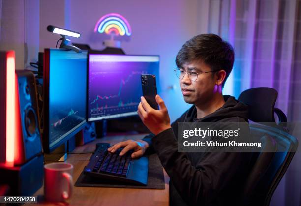 young asian male trading and currency trading app on his smartphone from his home office - cryptocurrency trading stock pictures, royalty-free photos & images