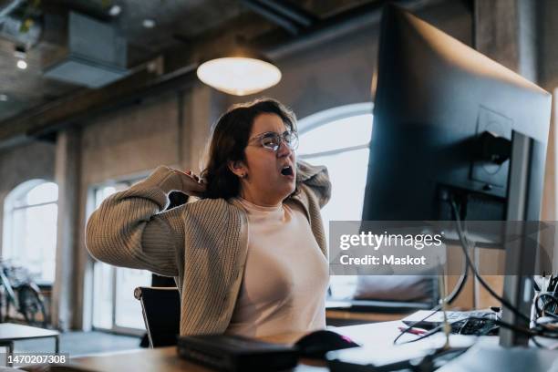 tired female hacker yawning and stretching hands at desk in creative office - gapen stockfoto's en -beelden