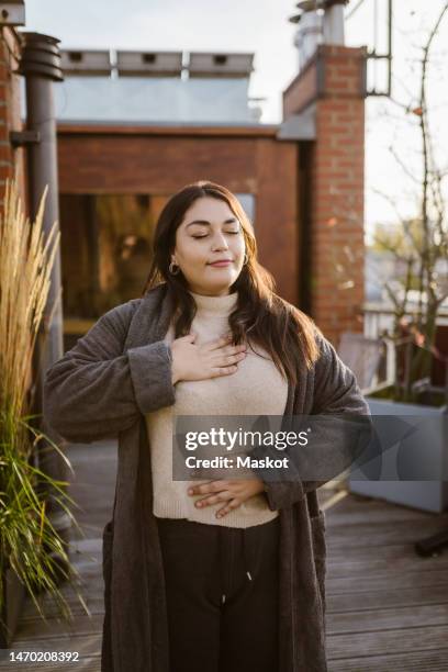 woman with eyes closed practicing breathing exercise while standing at terrace - hands on chest stock pictures, royalty-free photos & images