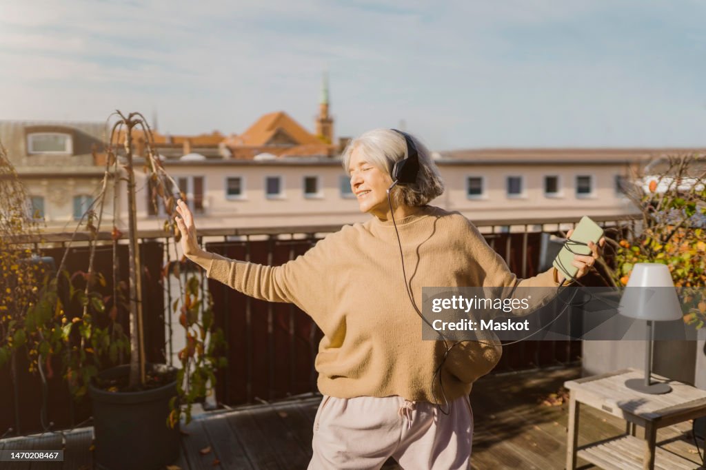 Carefree senior woman enjoying listening to music through headphones while standing at terrace