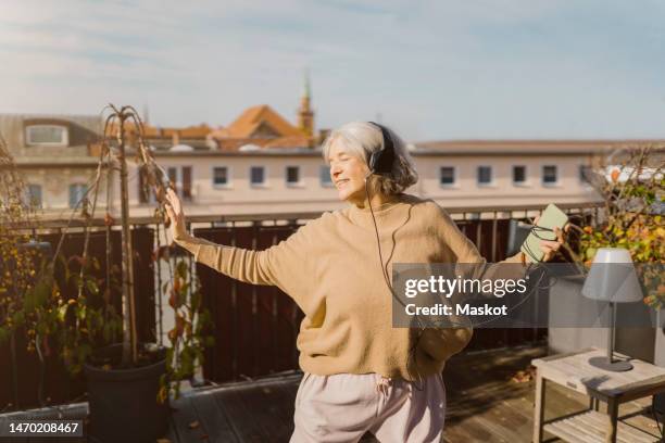 carefree senior woman enjoying listening to music through headphones while standing at terrace - alleen seniore vrouwen stockfoto's en -beelden