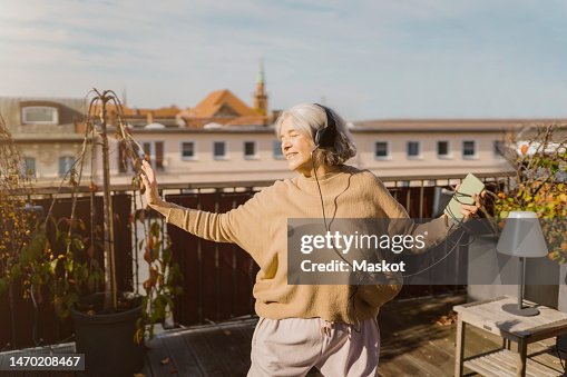 Carefree senior woman enjoying listening to music through headphones while standing at terrace Carefree senior woman enjoying listening to music through headphones while standing at terrace
