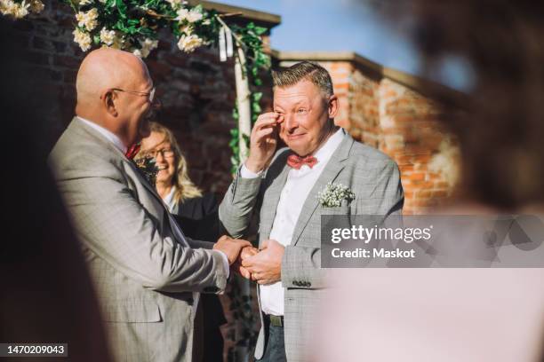 smiling groom wiping his tears with finger while holding hand of partner by minister on sunny day - hochzeitsgesellschaft stock-fotos und bilder
