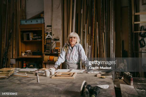 portrait of confident senior female carpenter leaning on workbench at repair shop - handgemacht stock-fotos und bilder