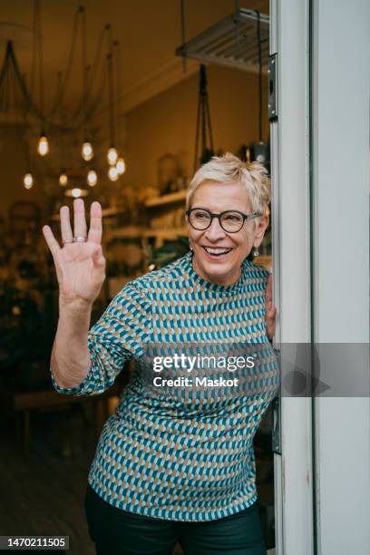 happy female entrepreneur waving while standing at doorway of store - wave hello outside stock pictures, royalty-free photos & images