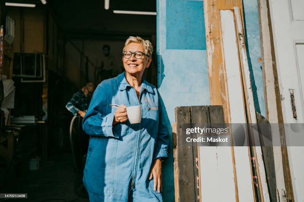 Happy female entrepreneur holding cup while standing near workshop doorway