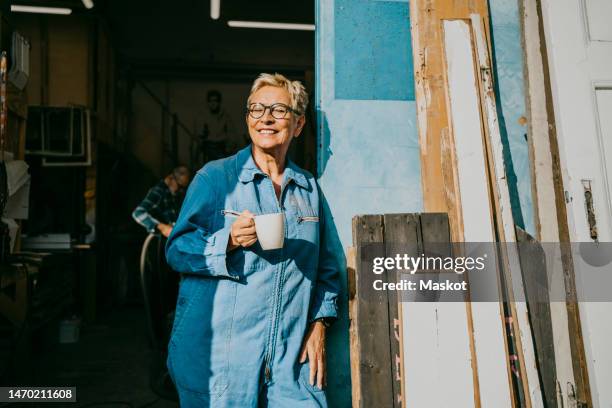 happy female entrepreneur holding cup while standing near workshop doorway - working seniors stock pictures, royalty-free photos & images