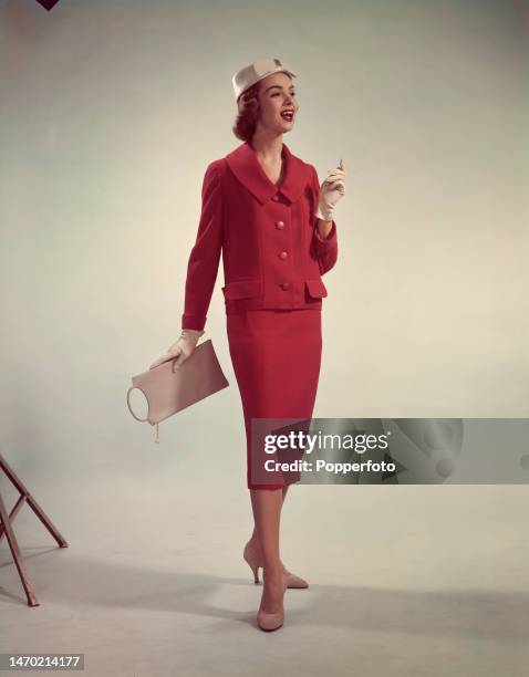Posed studio portrait of a woman wearing a two piece red spring suit comprising a short boxy jacket with tab pockets and a slim pencil skirt, it is...