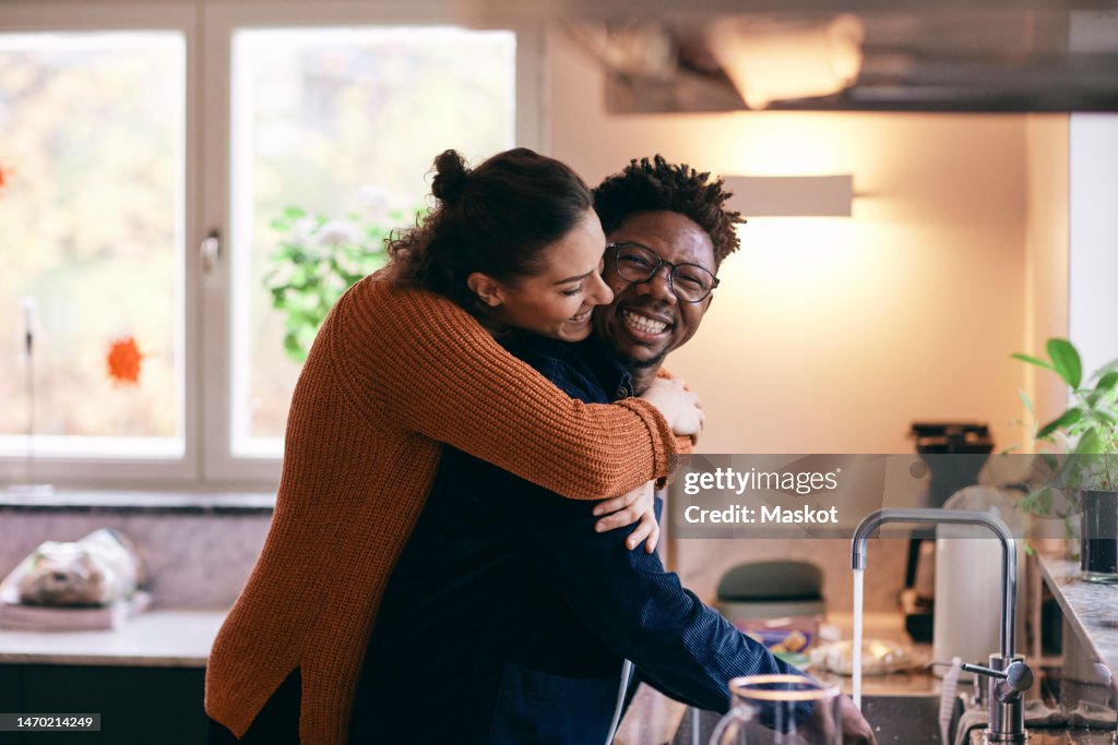 Side view of happy woman embracing man washing hands in kitchen at home