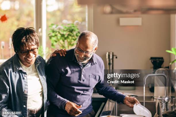 happy senior woman with hand on shoulder of man washing dishes in kitchen at home - ethnicity stock pictures, royalty-free photos & images