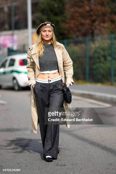 Guest wears a brown and black braided wool beret from Ganni, a white cropped shirt, a dark gray braided wool cropped pullover, a beige long trench...