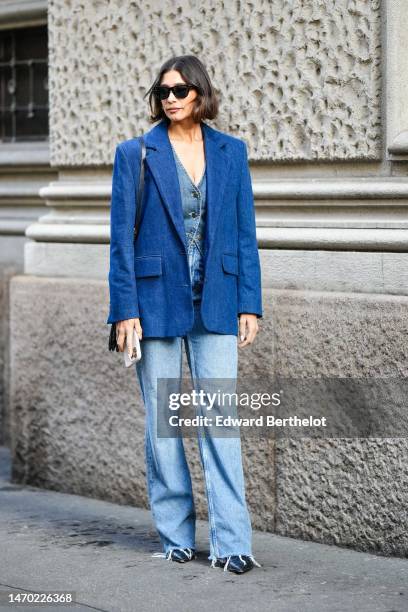 Guest wears black sunglasses, gold earrings, a blue V-neck / buttoned denim gilet, a blue denim blazer jacket, a black shiny leather shoulder bag,...