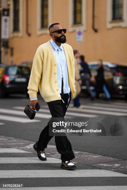 Guest wears black sunglasses, a white and baby blue striped print pattern shirt, a pale yellow buttoned wool cardigan, black suit pants, a black...