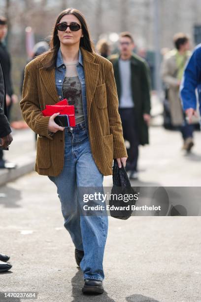 Guest wears black sunglasses, a beige with khaki and black print pattern tulle t-shirt, a blue denim shirt, a brown ribbed velvet oversized blazer...