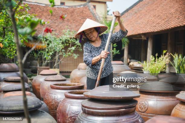 vietnamese senior woman stirring in huge clay soy pod in courtyard - asian style conical hat stock pictures, royalty-free photos & images
