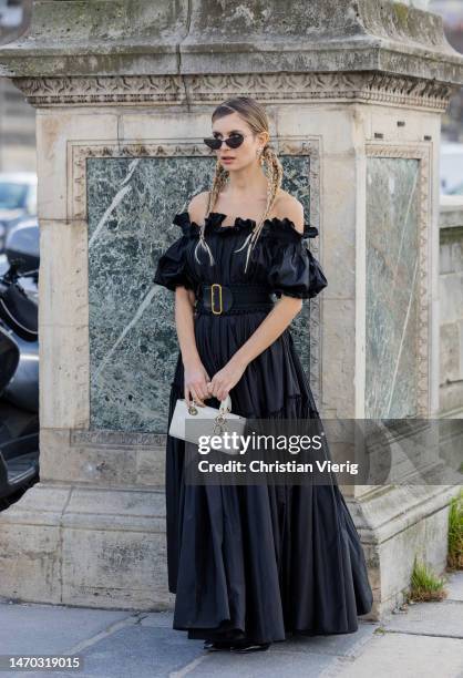 Xenia Adonts wears black off shoulder dress, belt, white bag outside Dior during Paris Fashion Week - Womenswear Fall Winter 2023 2024 : Day Two on...