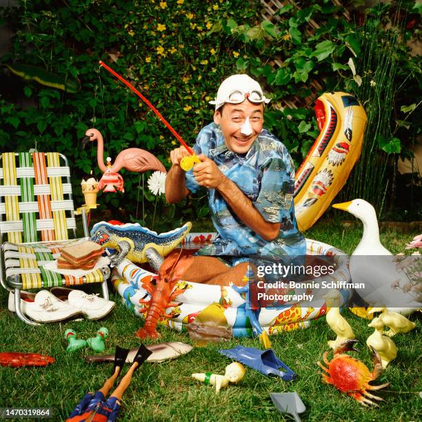 Pee-Wee Herman kneels in a kiddie pool, wears a swim cap with goggles, and fishes with a plastic rod amongst scattered toys and plastic fish in Los...