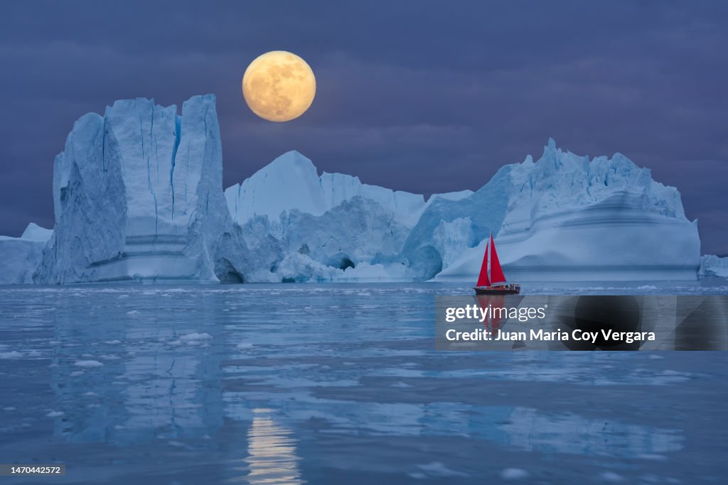 Red sailboat sailing among floating icebergs in front of the full moon rising at Arctic Ocean in Greenland, Ilulissat Icefjord, Ilulissat, Disko Bay, Unesco World Heritage Site