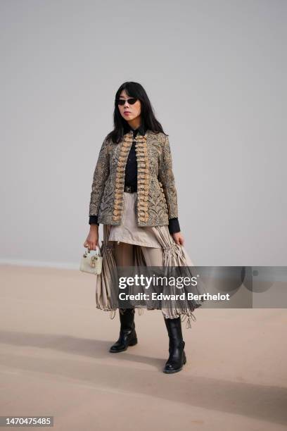 Guest wears black vintage sunglasses, a black shirt, a beige with embroidered black pattern / belted buttoned jacket from Dior, a black belt from...