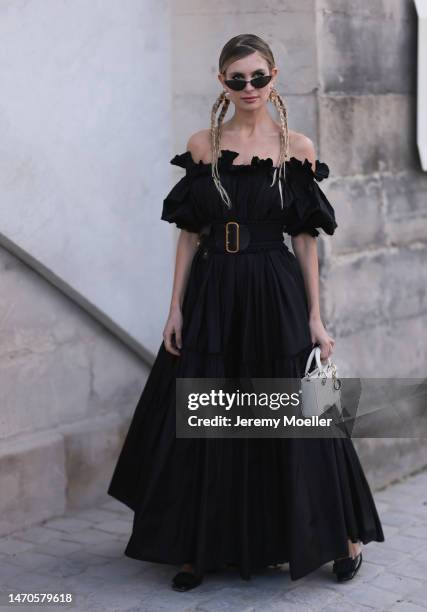 Xenia Adonts seen wearing a black shoulder free dress with a big black belt, black shades and a white bag before the Dior show on February 28, 2023...