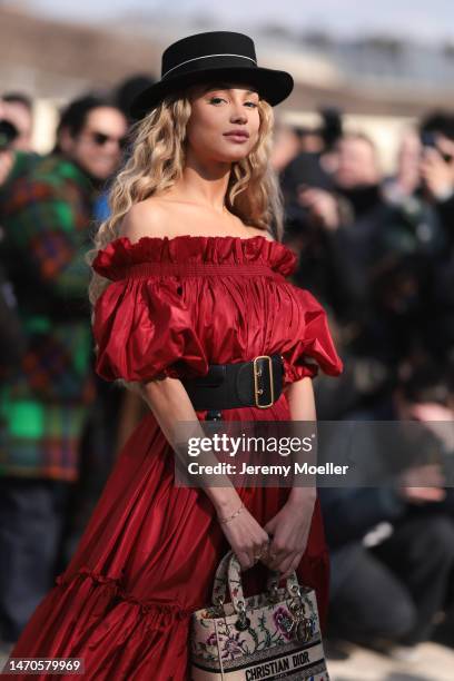 Rose Bertram seen wearing a red shoulder free dress, a big black belt, a black hat and a beige flower patterned bag before the Dior show on February...