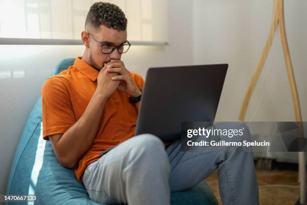 worried young man, sitting on a pouf, working on the laptop on his legs - latina legs stock-fotos und bilder