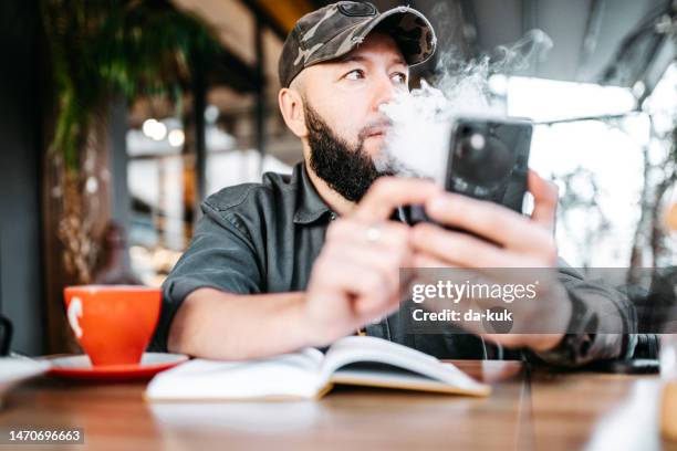 morning coffee and vape before work. casual man vaping sitting in cafe and using phone and note pad - café da internet stock pictures, royalty-free photos & images