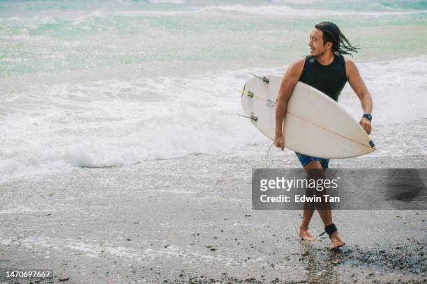 asian chinese male surfer walking on beach back from sea after riding wave in pacific ocean - asian surfer stockfoto's en -beelden