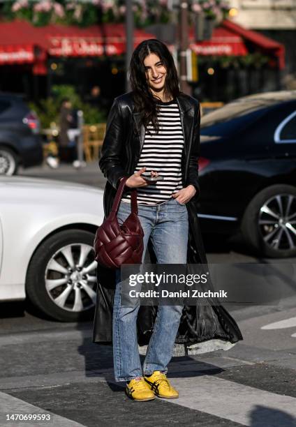 Model is seen wearing a black leather coat, black and white striped shirt, blue jeans and yellow sneakers with a maroon bag outside the Dries Van...