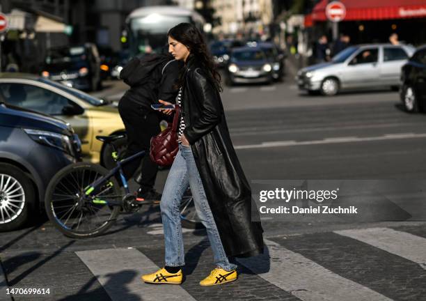 Model is seen wearing a black leather coat, black and white striped shirt, blue jeans and yellow sneakers with a maroon bag outside the Dries Van...