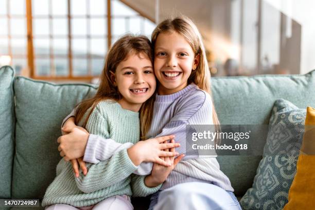 Portrait Of Two Little Sisters Sitting In Sofa At Home Embracing Each Other, Stockfoto