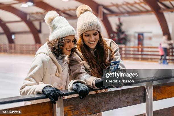 two female friends using phone while ice skating indoors - ice skate stock pictures, royalty-free photos & images