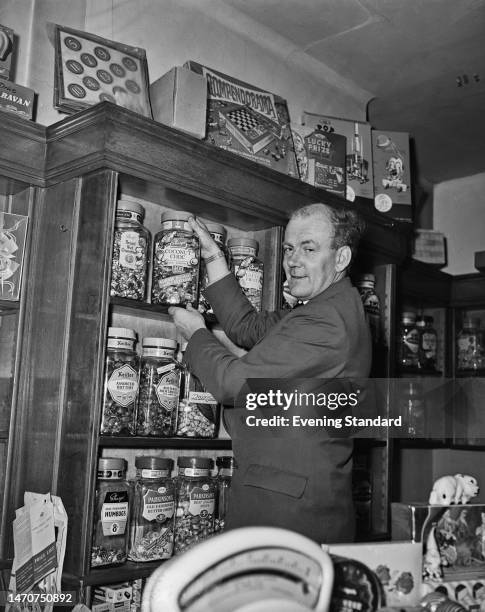 British cricketer Tony Lock takes a jar of coconut choc soft centre sweets from a shelf containing jars of sweets, United Kingdom, 3rd February 1961....