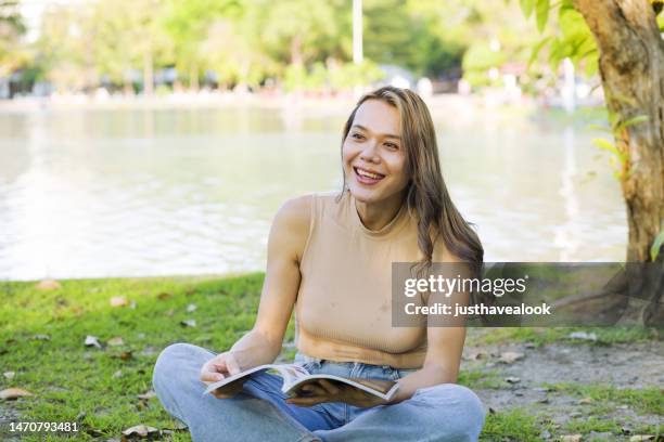 looking up and smiling   thai transgender sitting  with  book  in public park - kathoey stock pictures, royalty-free photos & images