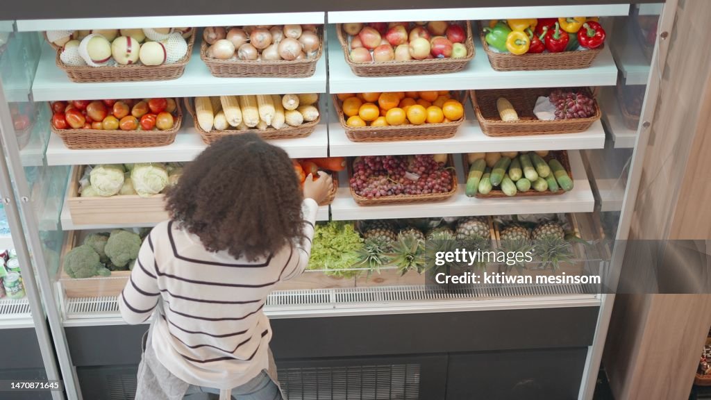Top view. African American worker in apron stocking the fruits in supermarket. Young employee at work, replenishes products on the shelves, works in a grocery store. Female arranging vegetable and fruits on shelf an organic store in shopping mall.