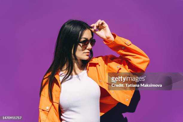 a beautiful young asian woman in sunglasses and a bright orange jacket on a purple background. - el milenio fotografías e imágenes de stock