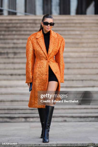 Guest wears black puffy sunglasses, gold earrings, a black turtleneck short tube dress, an orange with embroidered gold sequined long buttoned coat,...