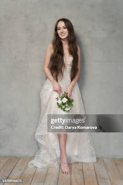 beautiful young woman with long brown hair in wedding dress without shoes and with flowers on grey background with copy space. the happy woman is smiling. the concept of tired feet during the wedding day. - female foot models fotografías e imágenes de stock