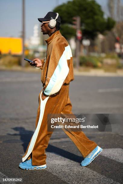 Guest wears a black with white embroidered logo pattern cap from New Era, beige sunglasses, gray headphones, black pearls in the hair, a brown velvet...