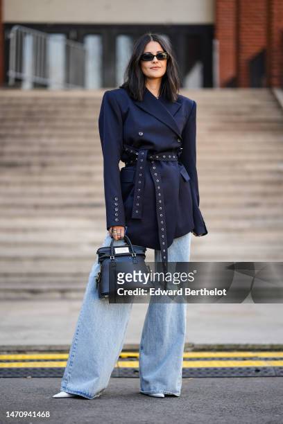 Guest wears black sunglasses, a navy blue oversized blazer jacket with belt nailed / studded from Off-White, pale blue denim wide legs pants, a black...