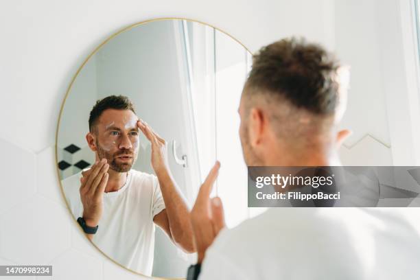 a millennial man is using a face cream in the bathroom - gezichtscrème stockfoto's en -beelden