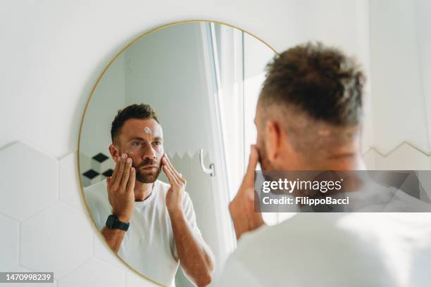 a millennial man is using a face cream in the bathroom - man eye cream imagens e fotografias de stock