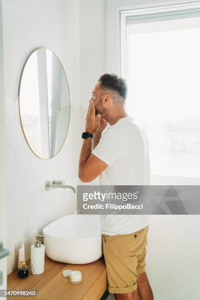 a millennial man is using a face cream in the bathroom - man eye cream imagens e fotografias de stock
